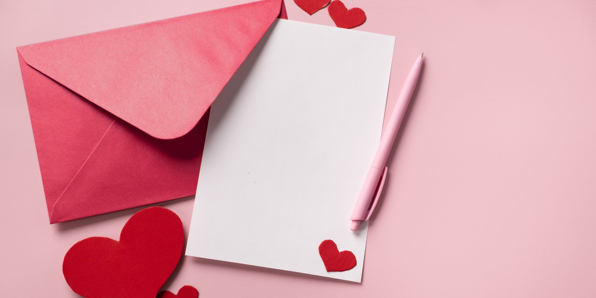 A pink envelope, a blank white card, a pink pen, and red heart decorations are arranged on a pink background.