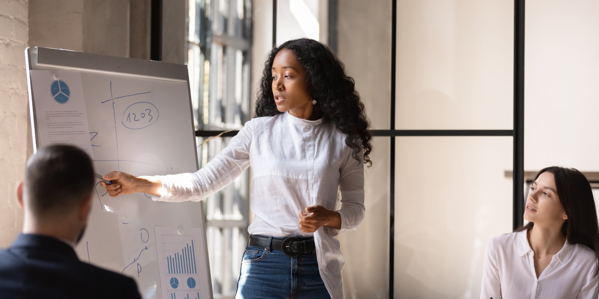 A woman is pointing at a whiteboard with charts and graphs, presenting to two colleagues who are seated and listening.