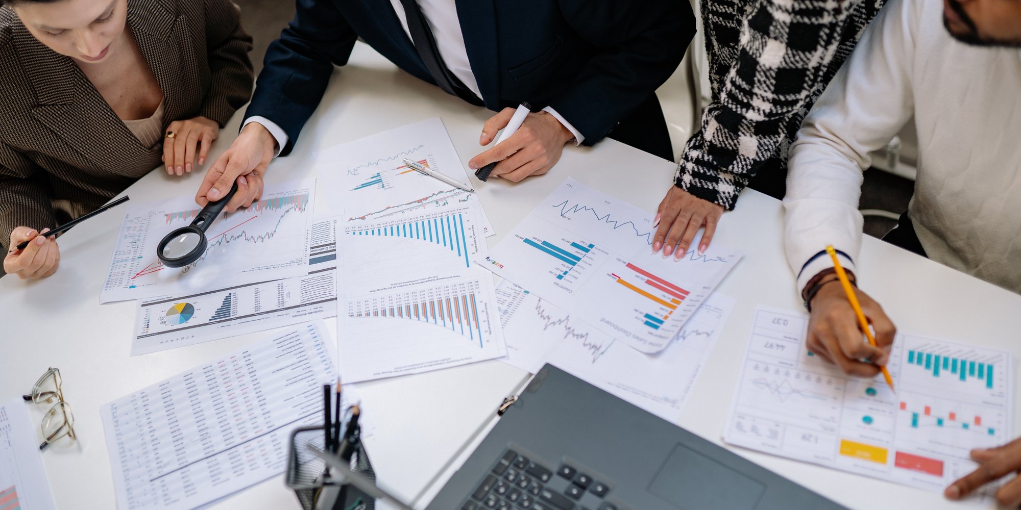 A group of people gathered around a table, analyzing charts and graphs with a magnifying glass and pens.