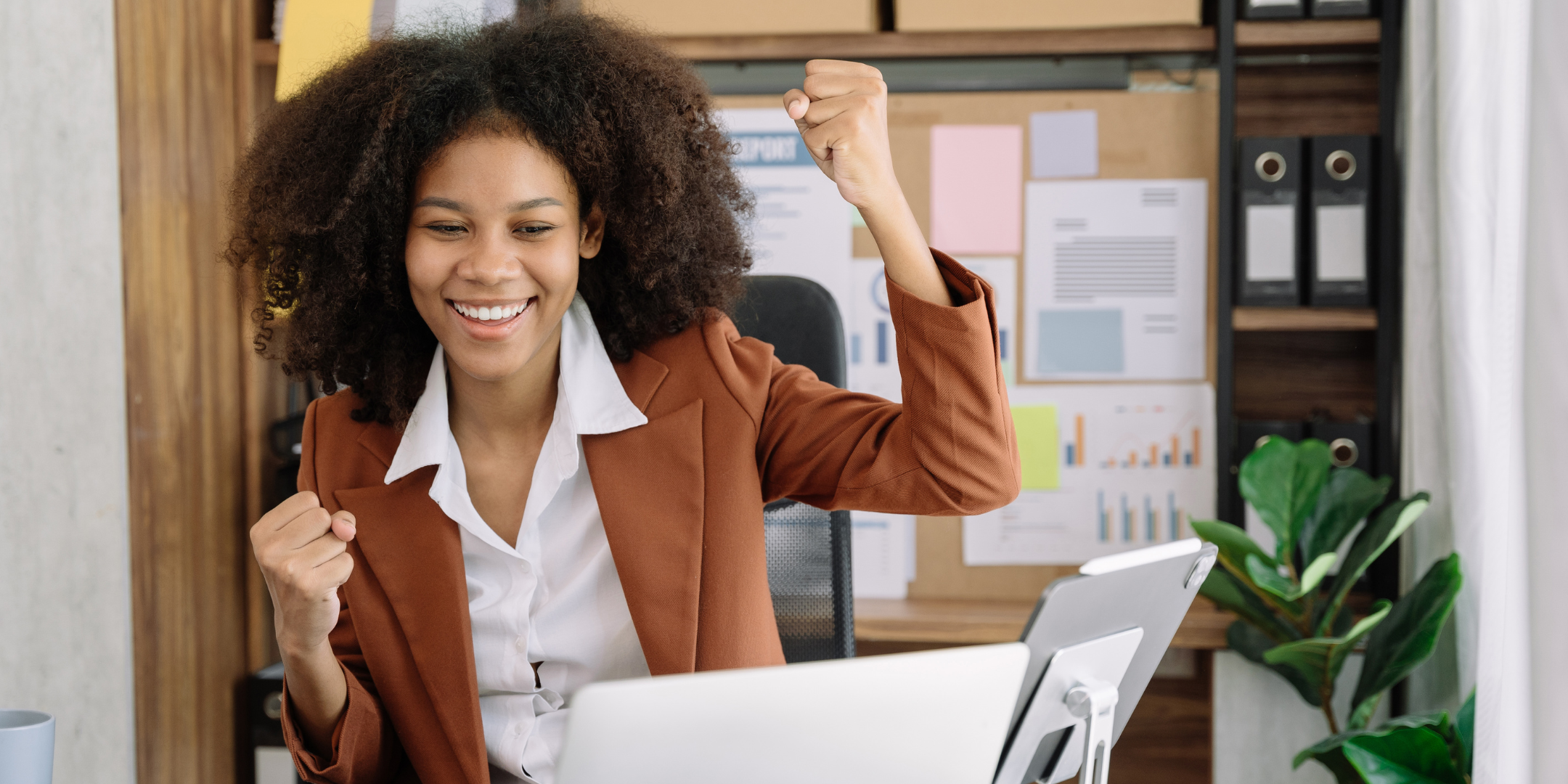 A person in a brown blazer sits at a desk with a laptop and tablet, raising their fists in celebration.