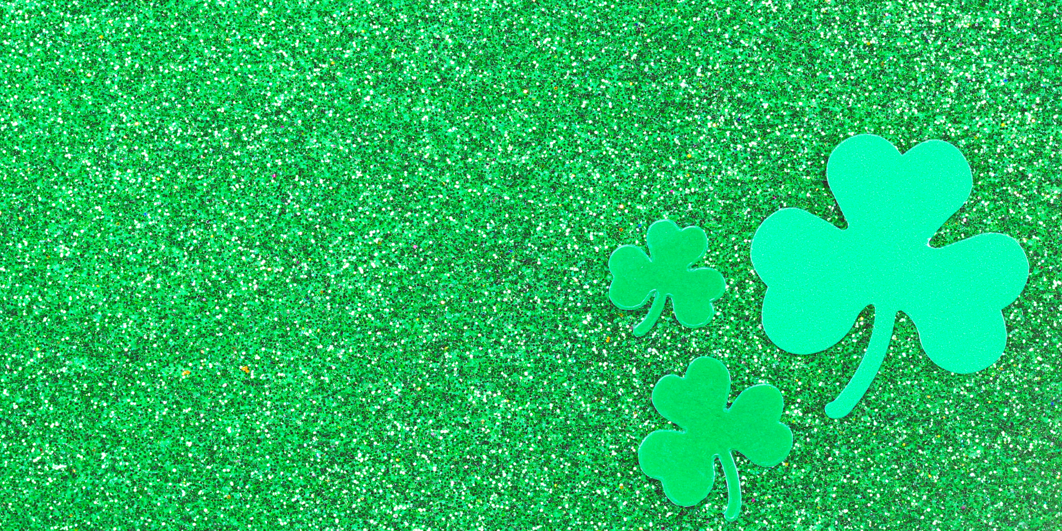 Three green shamrocks are placed on a sparkly green background. The largest shamrock is on the right, with two smaller shamrocks below and to its left. The background has a textured, glittering appearance.