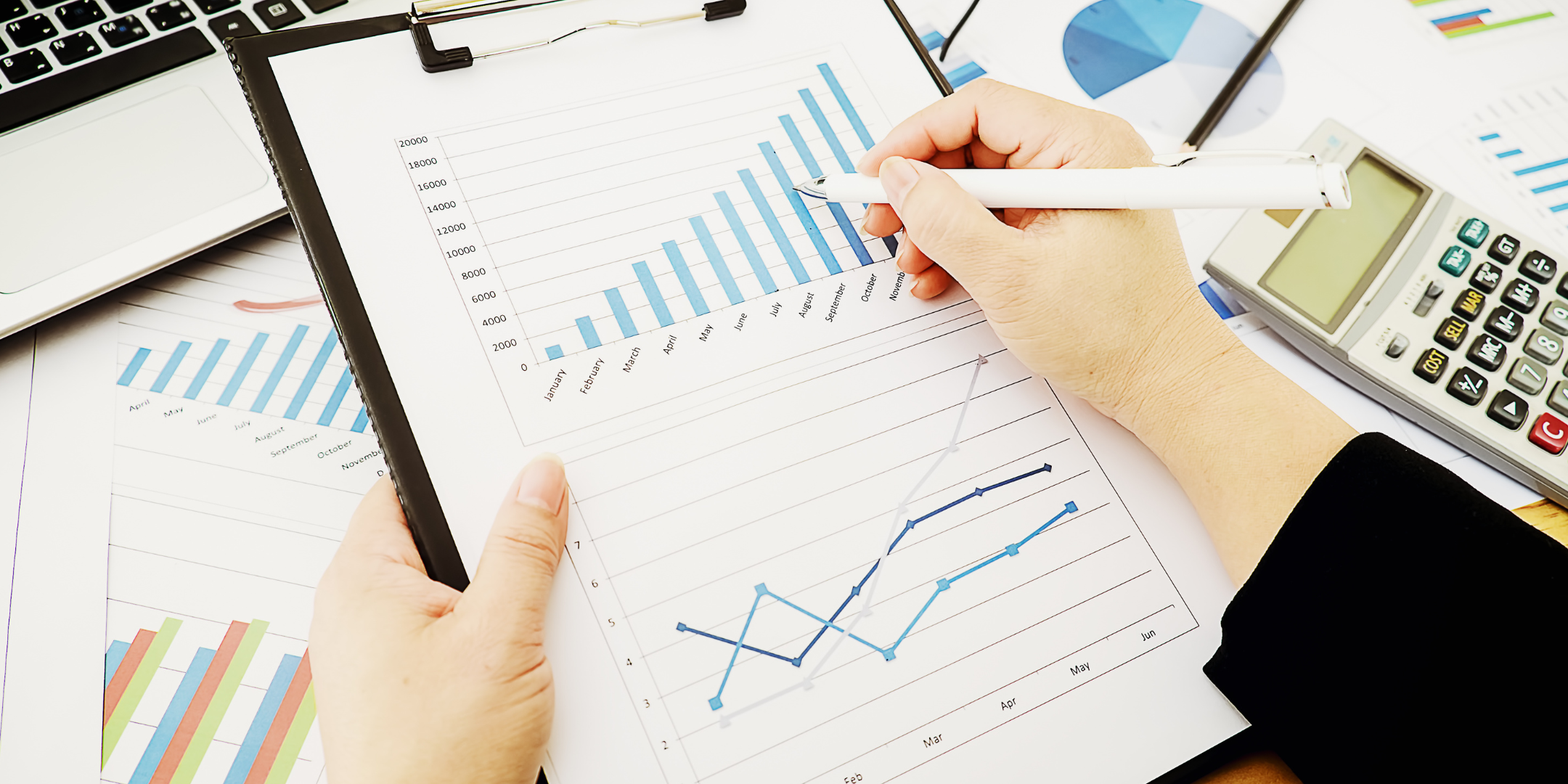 Hands holding a clipboard with financial charts, with a laptop and calculator nearby.