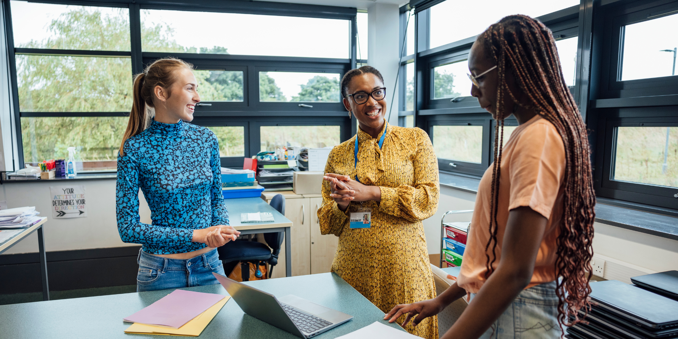 A teacher wearing glasses and a yellow dress stands between two students in a classroom, smiling and gesturing. One student wears a blue patterned top and jeans, the other a peach t-shirt and shorts.